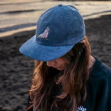 Woman wearing a blue coduroy hat with a northern elephant seal embroidered on it  on a beach at sunset
