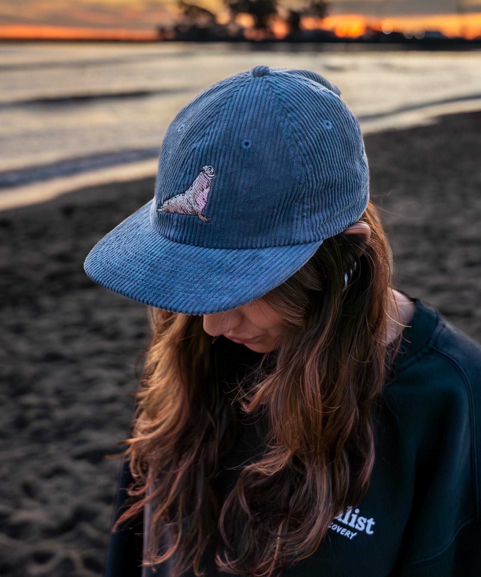 Woman wearing a blue coduroy hat with a northern elephant seal embroidered on it  on a beach at sunset
