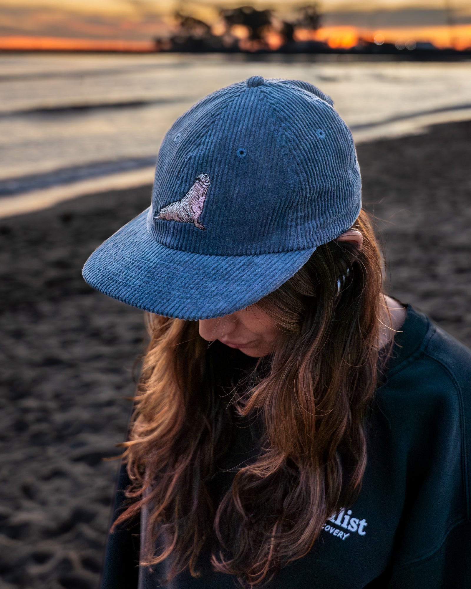 Woman wearing a blue coduroy hat with a northern elephant seal embroidered on it  on a beach at sunset