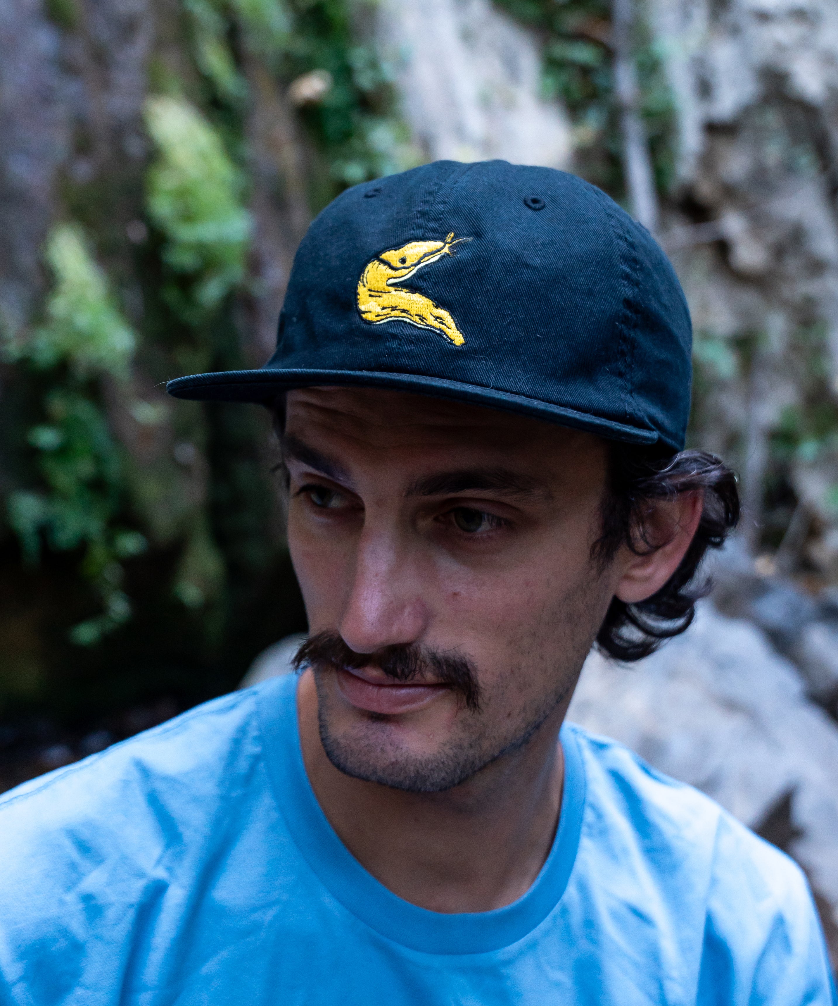 Man wearing a navy cap with a yellow banana slug in front of a waterfall