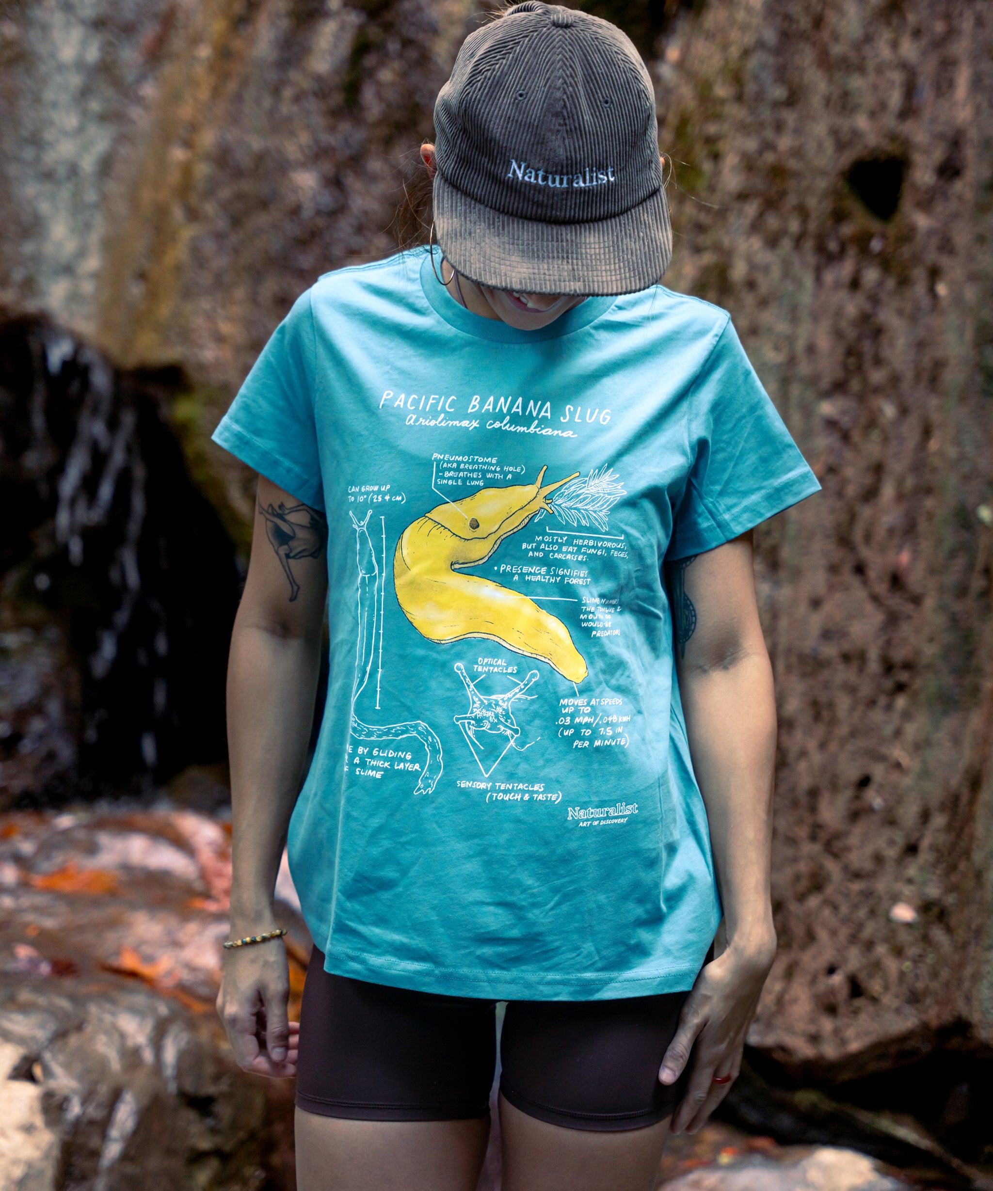 Person wearing a blue t-shirt with a yellow banana graphic and a cap, standing against a rocky background.