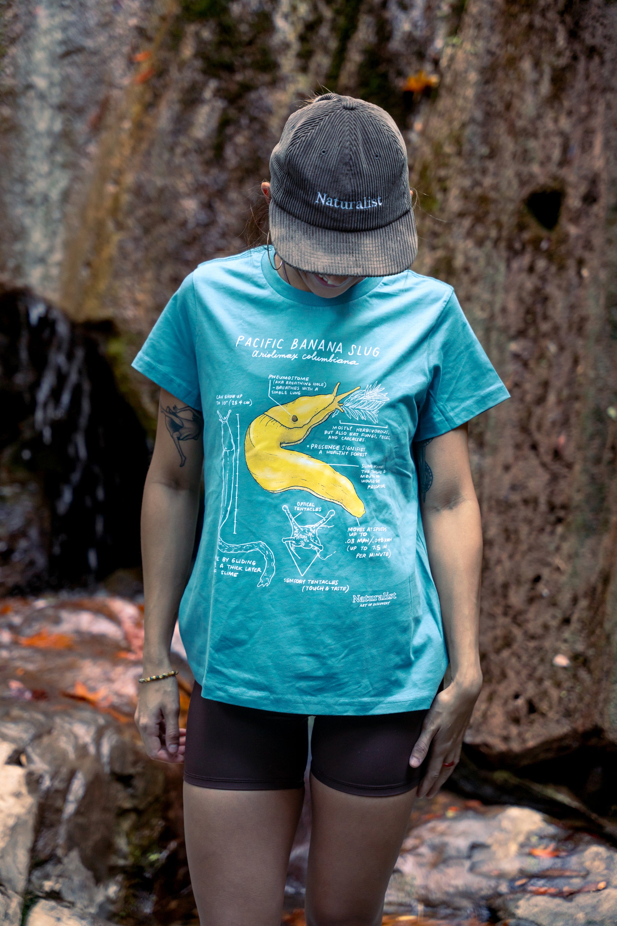 Person wearing a blue t-shirt with a yellow banana graphic and a cap, standing against a rocky background.