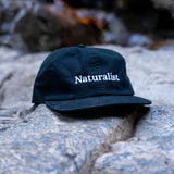 Black cap with 'Naturalist' text on a rock surface