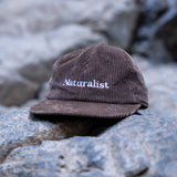 Brown corduroy cap with 'Naturalist' text on a rocky surface