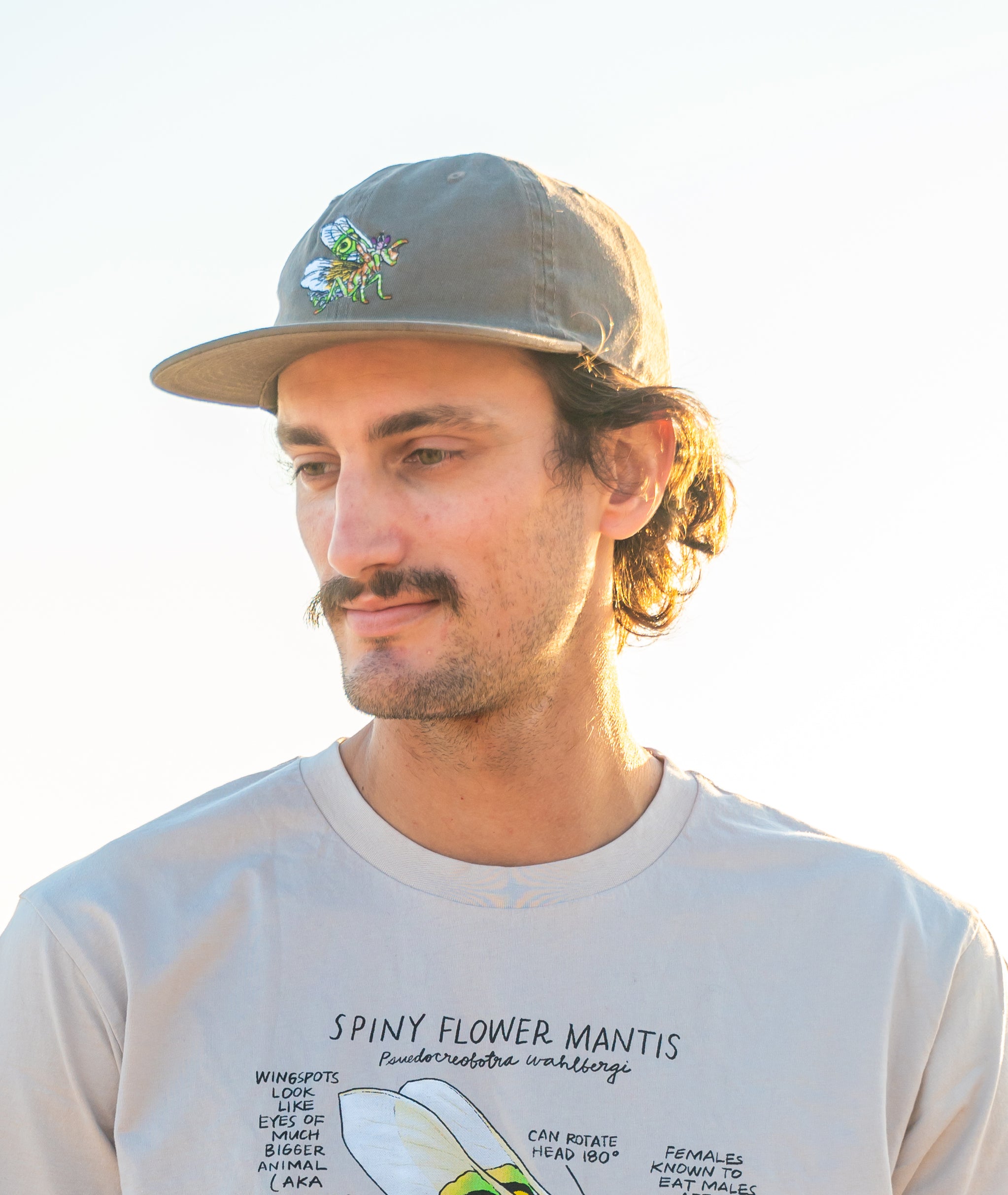 Man wearing a cap and t-shirt with text an illustration of a spiny flower mantis, standing outdoors.