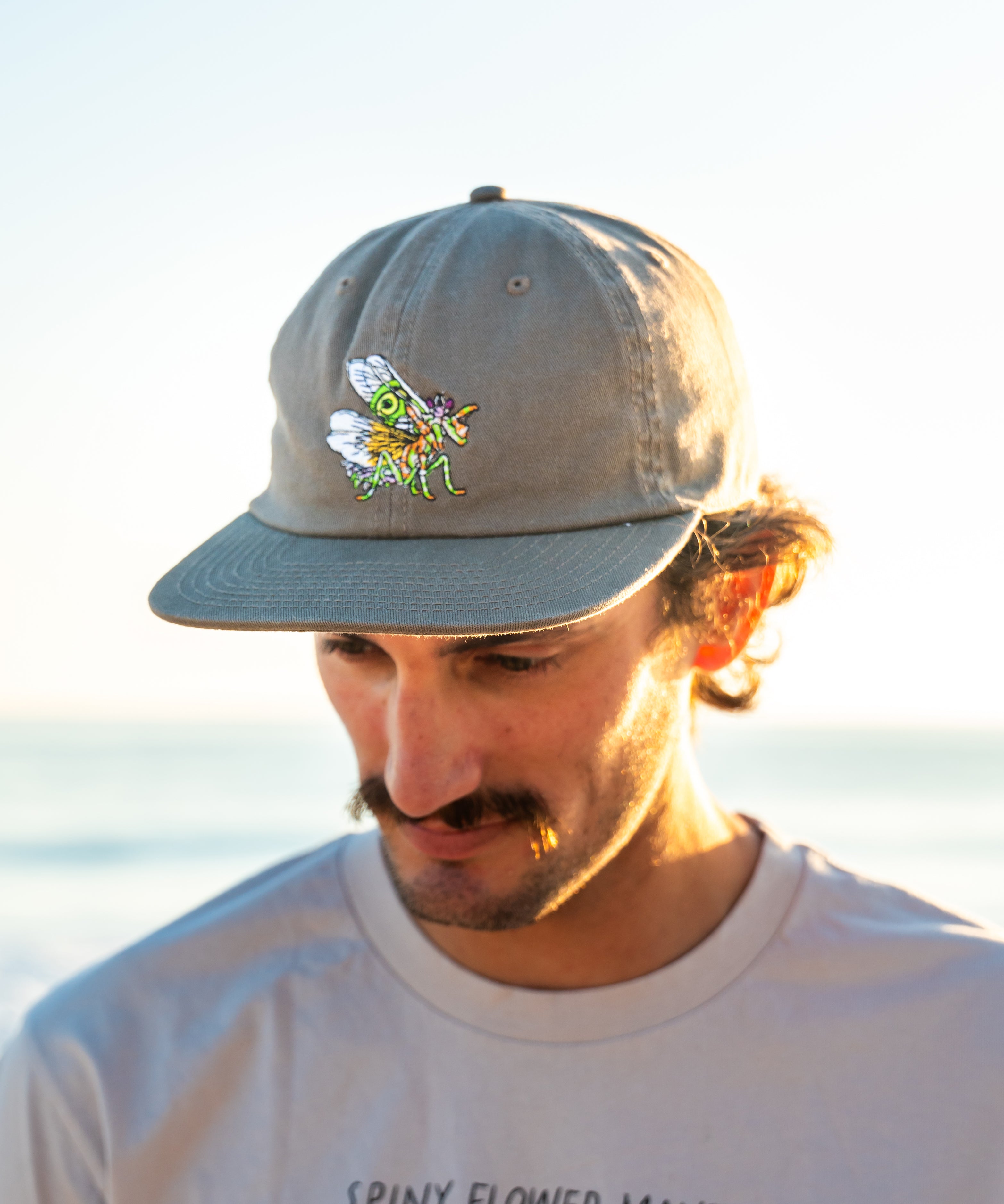Man wearing a cap with a spiny flower mantis standing by the ocean.
