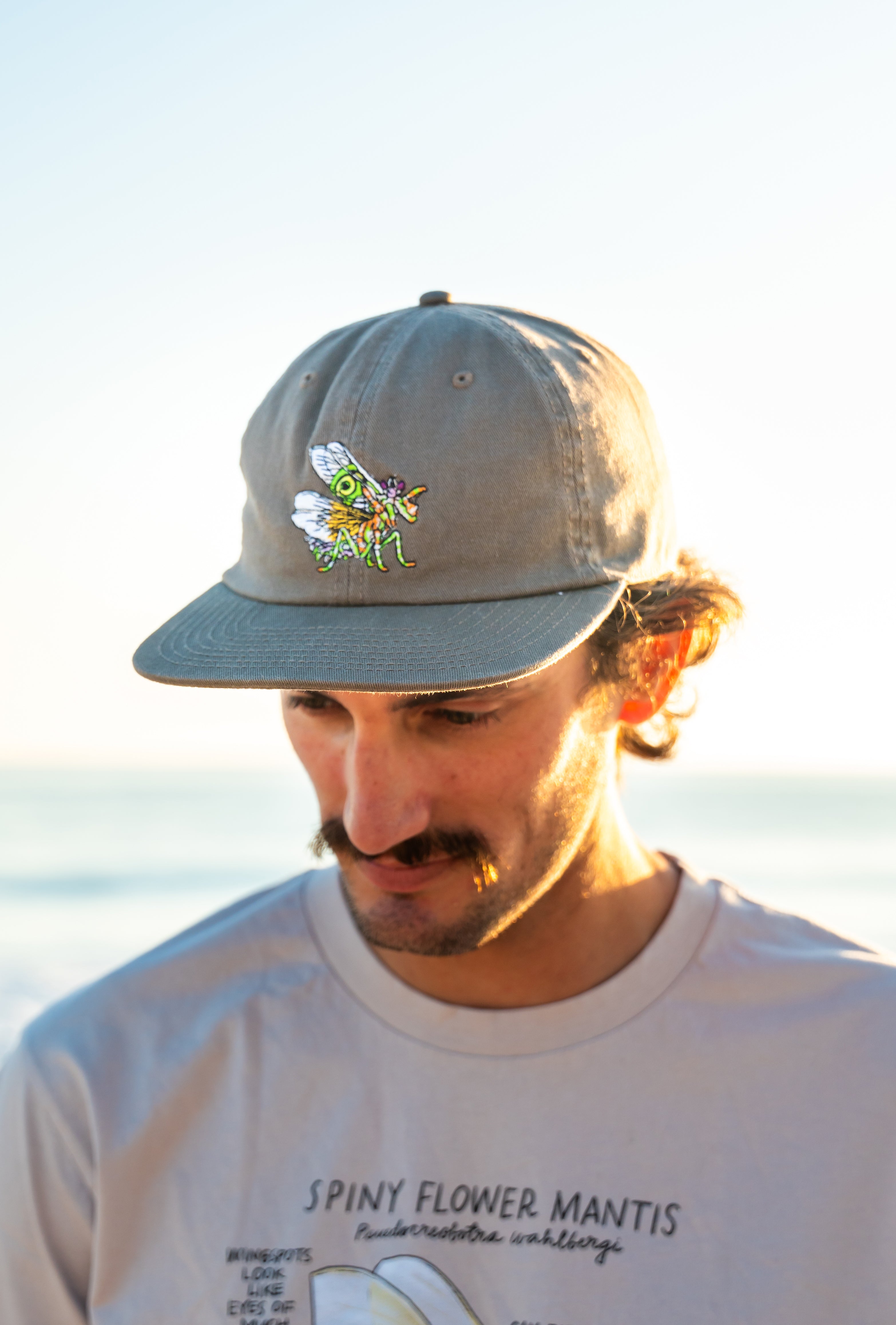 Man wearing a cap with a spiny flower mantis standing by the ocean.